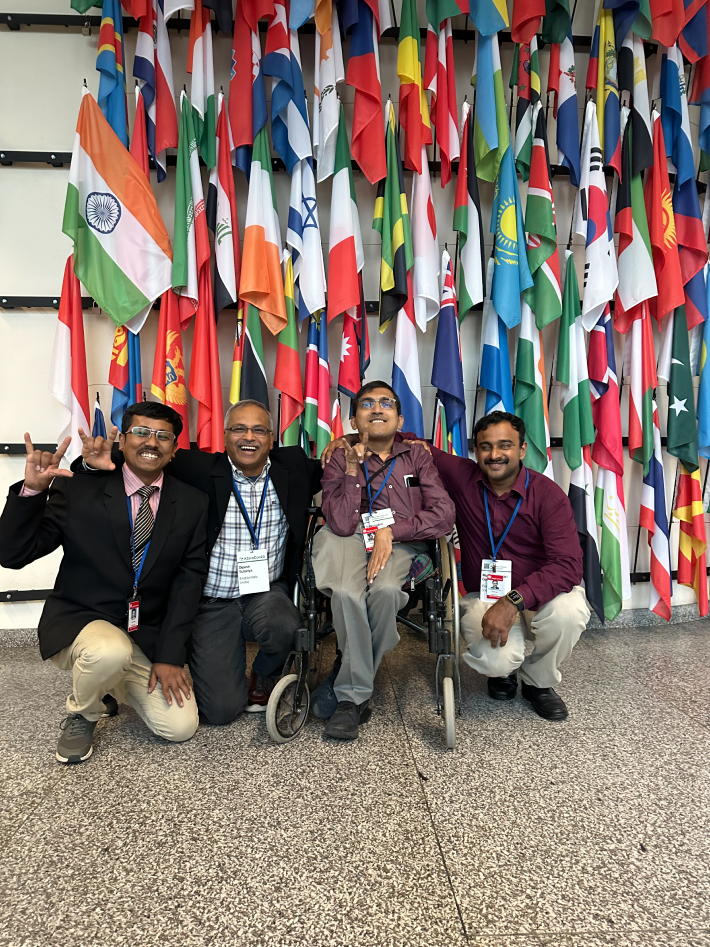 Four men, including two wheelchair users, smiling in front of international flags at an event celebrating disability innovation.