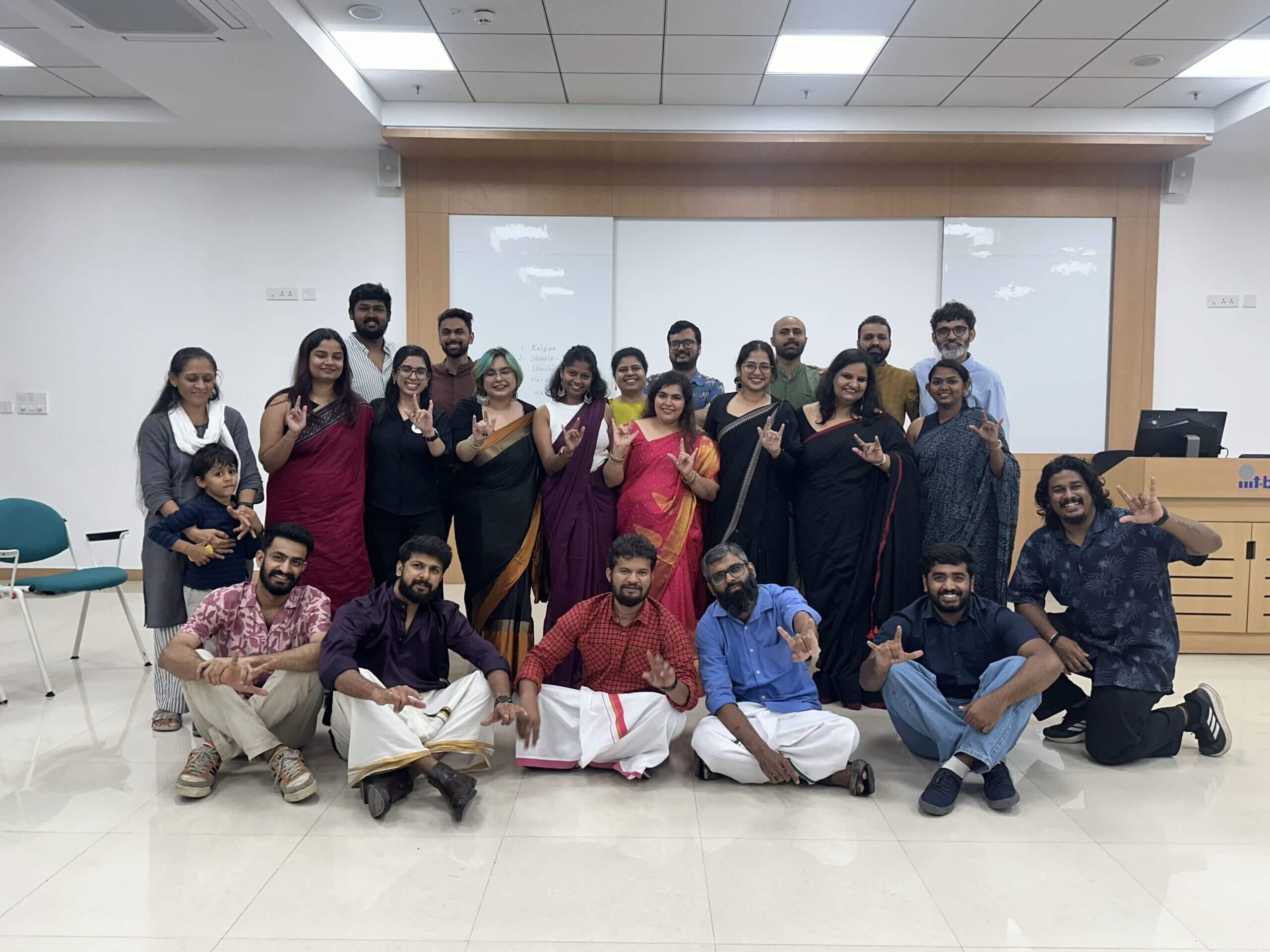 Group of twenty-one adults and one child in a classroom, with many making a hand gesture.