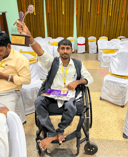 Akula Kumar, a man in a wheelchair, smiling and holding a trophy above his head in an event hall.