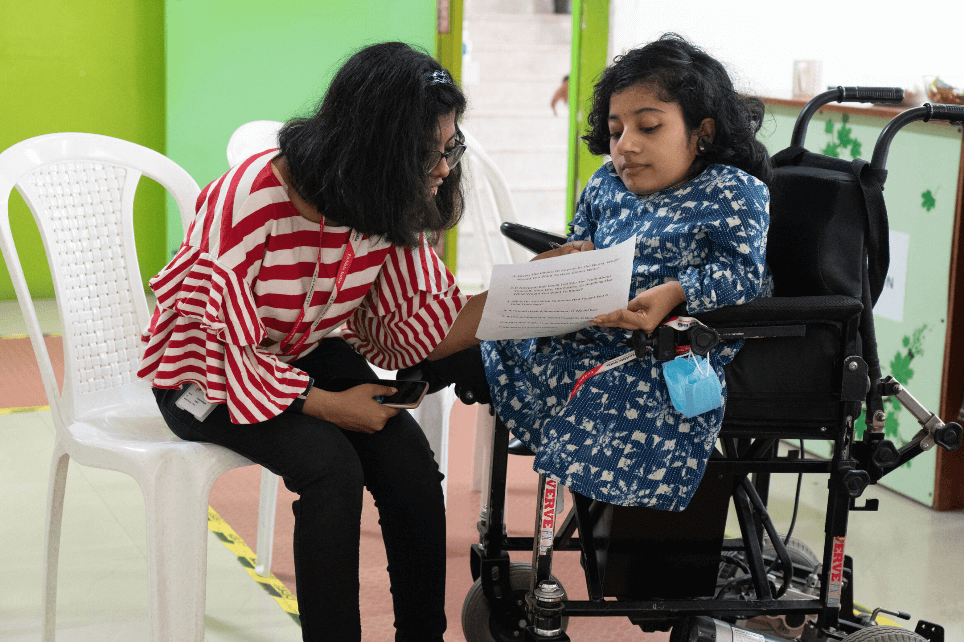 A young woman in a wheelchair reading a paper while another woman leans in to assist her.
