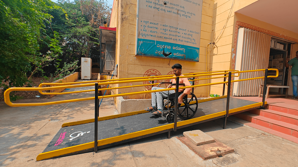 A man in a wheelchair using a bright yellow ramp installed outside a government building, part of the Ramp My City initiative.