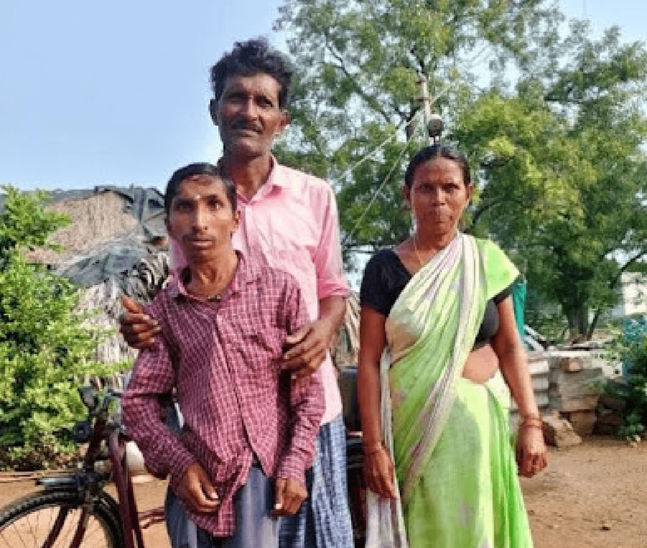 Boda Prasad standing with his parents in a rural setting, with a tricycle visible behind him