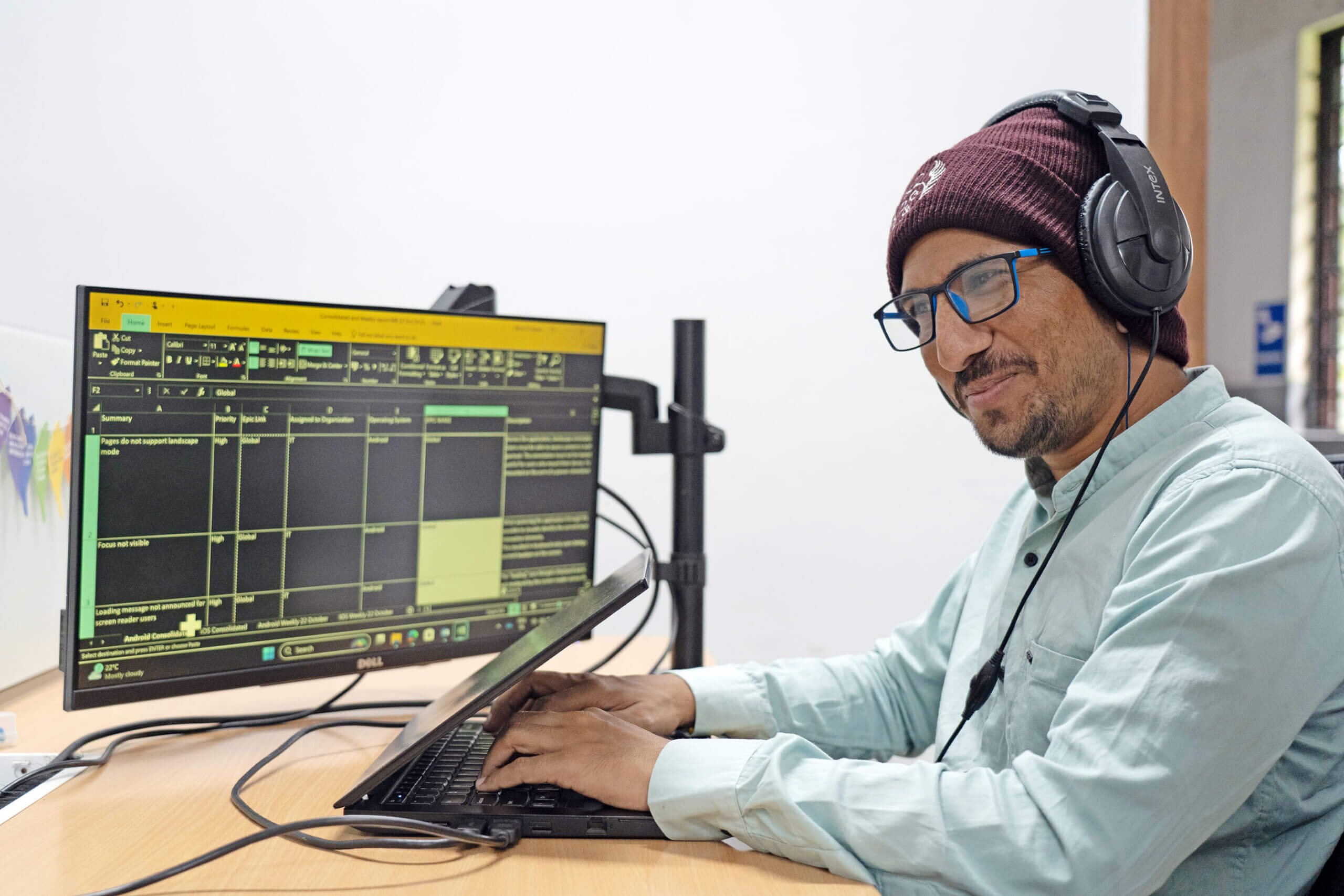 Smiling man wearing headphones and glasses working on a laptop with a dual monitor displaying code or accessibility testing data.
