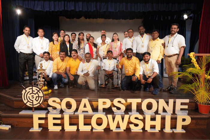 Group of men and women on a stage, posing behind a large "SOAPSTONE FELLOWSHIP" sign.